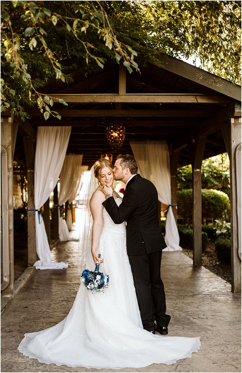 Premier Snohomish & Seattle Wedding Photographer | GSquared Weddings Photography A bride in a white gown holds a blue and white bouquet while a groom in a black suit kisses her cheek. They stand under a wooden pergola draped with white fabric, surrounded by greenery and soft sunlight at their Snohomish wedding. 47.9129° N, 122.0982° W | Serving Snohomish, Seattle , Orlando & Beyond