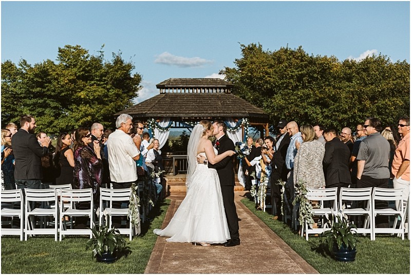 Premier Snohomish & Seattle Wedding Photographer | GSquared Weddings Photography A bride and groom kiss at the end of an outdoor aisle, surrounded by cheering guests. The wedding ceremony in Snohomish takes place before a gazebo beneath a clear, blue sky—capturing the charm of a Seattle Wedding. 47.9129° N, 122.0982° W | Serving Snohomish, Seattle , Orlando & Beyond