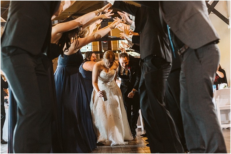 Premier Snohomish & Seattle Wedding Photographer | GSquared Weddings Photography A bride and groom walk under an arch of raised arms formed by their wedding party, smiling and celebrating as they enter a Snohomish Seattle wedding reception. The guests wear formal attire and the mood is joyful. 47.9129° N, 122.0982° W | Serving Snohomish, Seattle , Orlando & Beyond