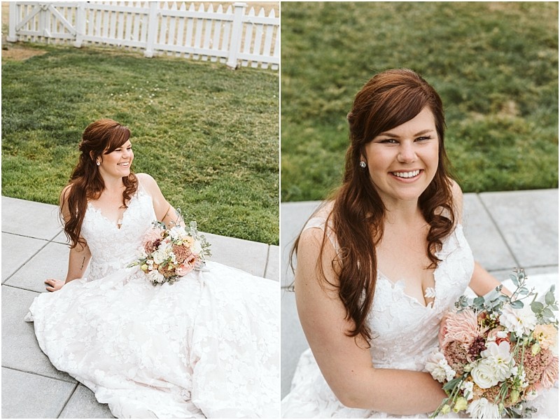 Premier Snohomish & Seattle Wedding Photographer | GSquared Weddings Photography A bride with long brown hair in a white lace wedding dress sits outdoors on a stone patio, holding a bouquet and smiling brightly. The scene captures the charm of a Snohomish Seattle wedding, with a grassy lawn and white picket fence behind her. 47.9129° N, 122.0982° W | Serving Snohomish, Seattle , Orlando & Beyond