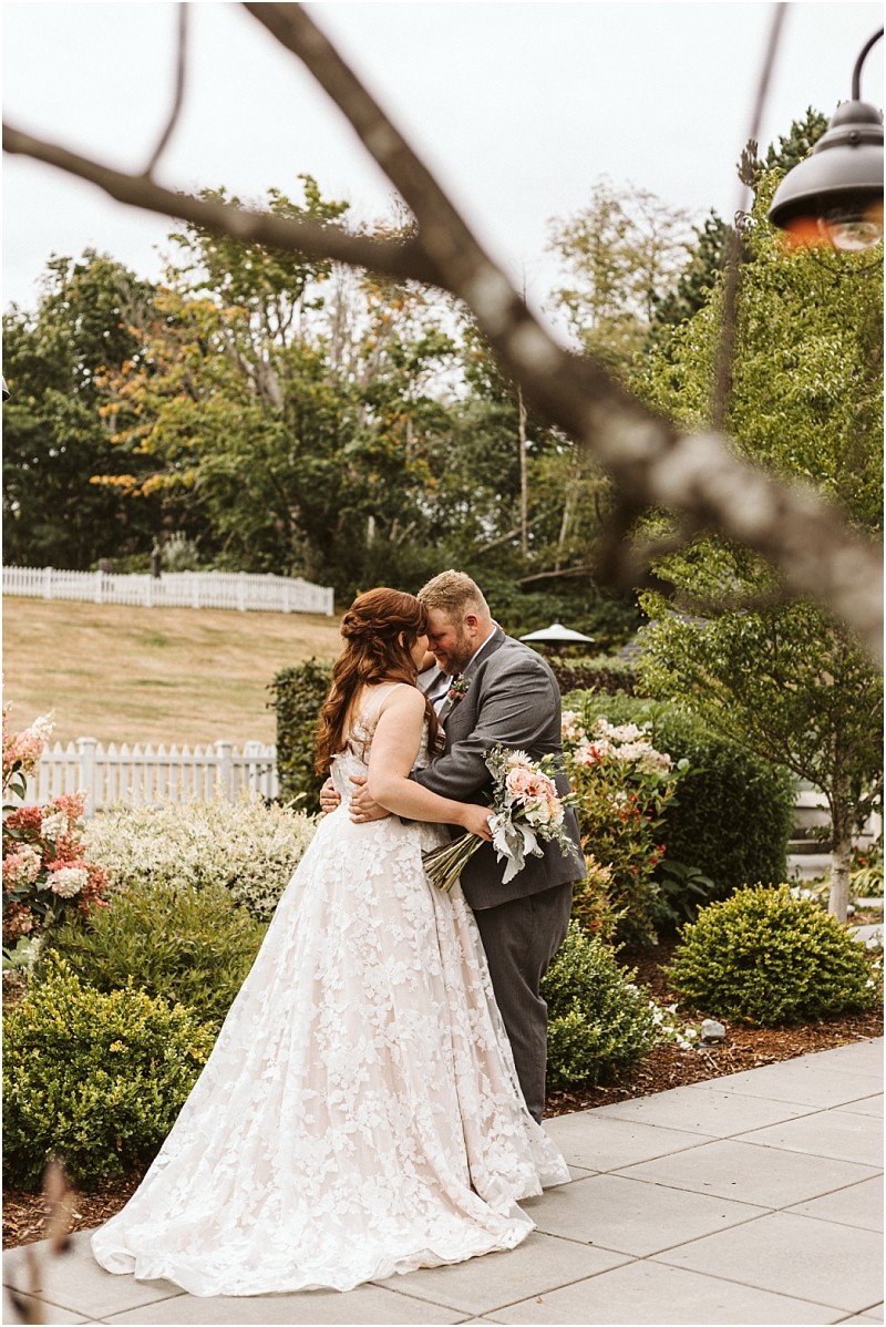 Premier Snohomish & Seattle Wedding Photographer | GSquared Weddings Photography A bride and groom embrace outdoors on a paved patio surrounded by lush greenery and flowers at their Snohomish wedding. The bride wears a white lace gown and holds a bouquet, while the groom wears a gray suit. Trees and a white fence are in the background. 47.9129° N, 122.0982° W | Serving Snohomish, Seattle , Orlando & Beyond