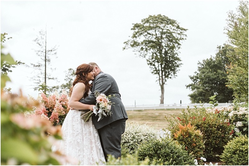 Premier Snohomish & Seattle Wedding Photographer | GSquared Weddings Photography A bride and groom embrace in a garden surrounded by flowers and greenery during their Snohomish wedding, with a large tree and open field in the background on a cloudy day. 47.9129° N, 122.0982° W | Serving Snohomish, Seattle , Orlando & Beyond