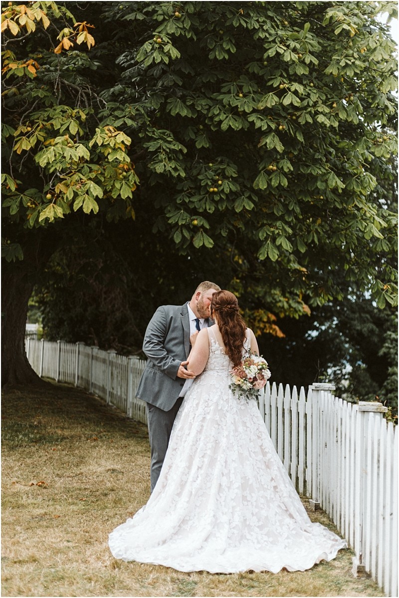 Premier Snohomish & Seattle Wedding Photographer | GSquared Weddings Photography A bride and groom share a kiss outdoors by a white picket fence at their Snohomish wedding, with the bride in a lace gown holding a bouquet and the groom in a gray suit. Large green trees provide shade in the background. 47.9129° N, 122.0982° W | Serving Snohomish, Seattle , Orlando & Beyond