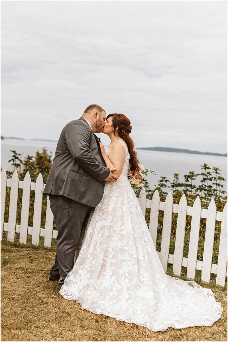 Premier Snohomish & Seattle Wedding Photographer | GSquared Weddings Photography A bride and groom share a kiss outdoors by a white picket fence, with water and greenery in the background during their Snohomish wedding. The bride wears a lace gown, while the groom is in a gray suit. 47.9129° N, 122.0982° W | Serving Snohomish, Seattle , Orlando & Beyond