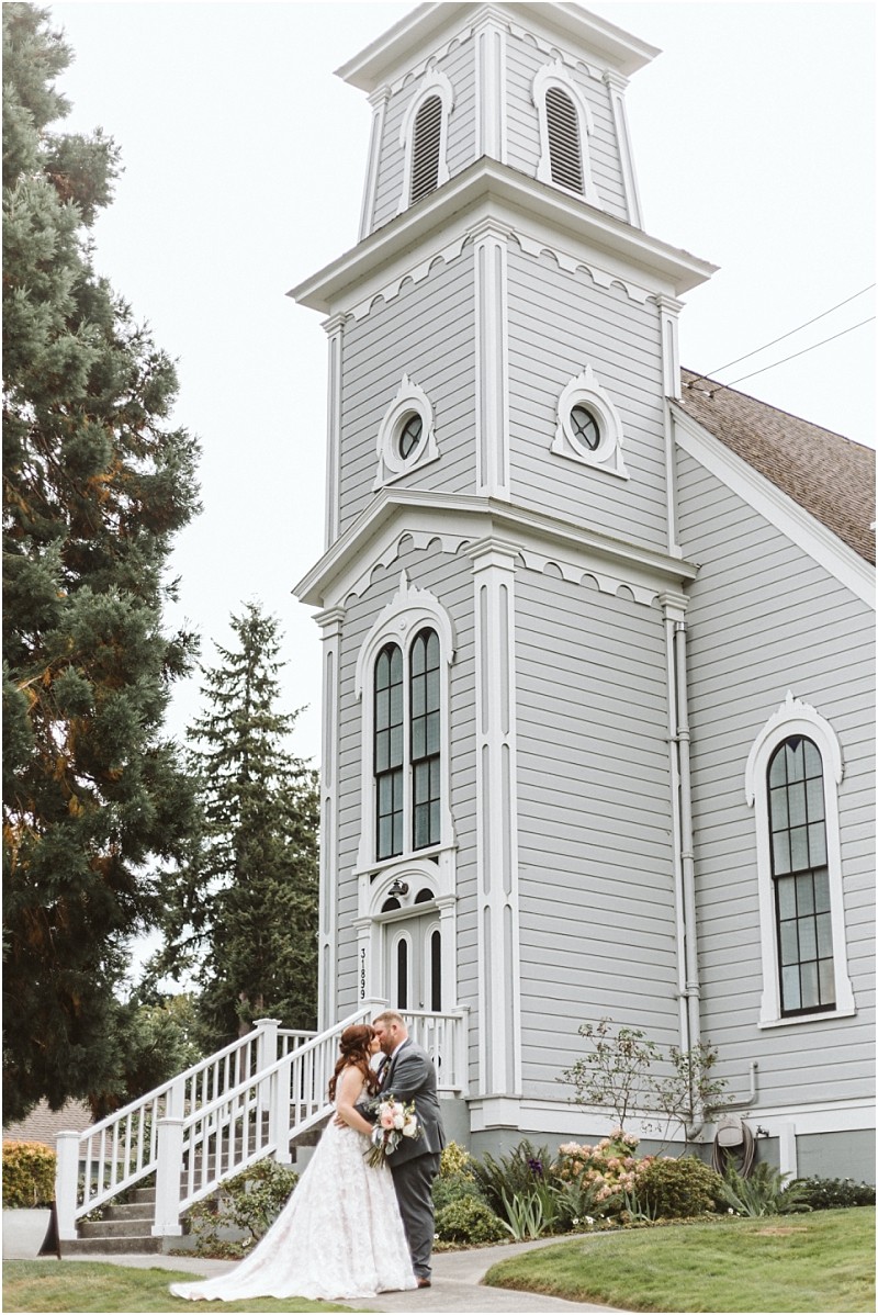 Premier Snohomish & Seattle Wedding Photographer | GSquared Weddings Photography A bride and groom stand together, looking at each other, outside a tall, light gray wooden church with large arched windows and white trim in Snohomish, surrounded by greenery on a cloudy Seattle wedding day. 47.9129° N, 122.0982° W | Serving Snohomish, Seattle , Orlando & Beyond