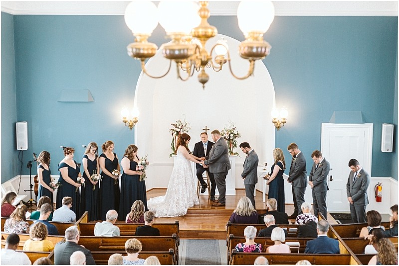 Premier Snohomish & Seattle Wedding Photographer | GSquared Weddings Photography A bride and groom stand at the altar in a church near Snohomish, surrounded by bridesmaids in navy dresses and groomsmen in gray suits, while guests watch the wedding ceremony from wooden pews. 47.9129° N, 122.0982° W | Serving Snohomish, Seattle , Orlando & Beyond