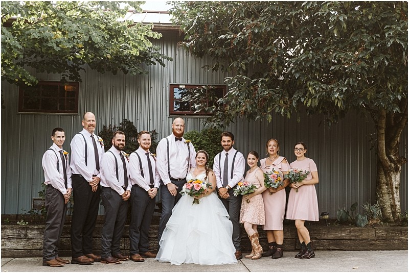 Premier Snohomish & Seattle Wedding Photographer | GSquared Weddings Photography A wedding party poses outdoors in front of a metal building at a Snohomish Seattle wedding. The bride in a white gown is surrounded by smiling bridesmaids in pink and groomsmen with suspenders, all holding bouquets and ready to celebrate the special day. 47.9129° N, 122.0982° W | Serving Snohomish, Seattle , Orlando & Beyond