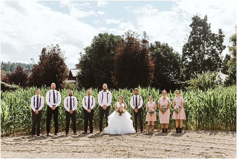 Premier Snohomish & Seattle Wedding Photographer | GSquared Weddings Photography A wedding party poses outdoors in front of tall green plants and trees at a beautiful Snohomish Seattle wedding. The bride stands in the center in a white dress, surrounded by bridesmaids in pink and groomsmen with suspenders. 47.9129° N, 122.0982° W | Serving Snohomish, Seattle , Orlando & Beyond