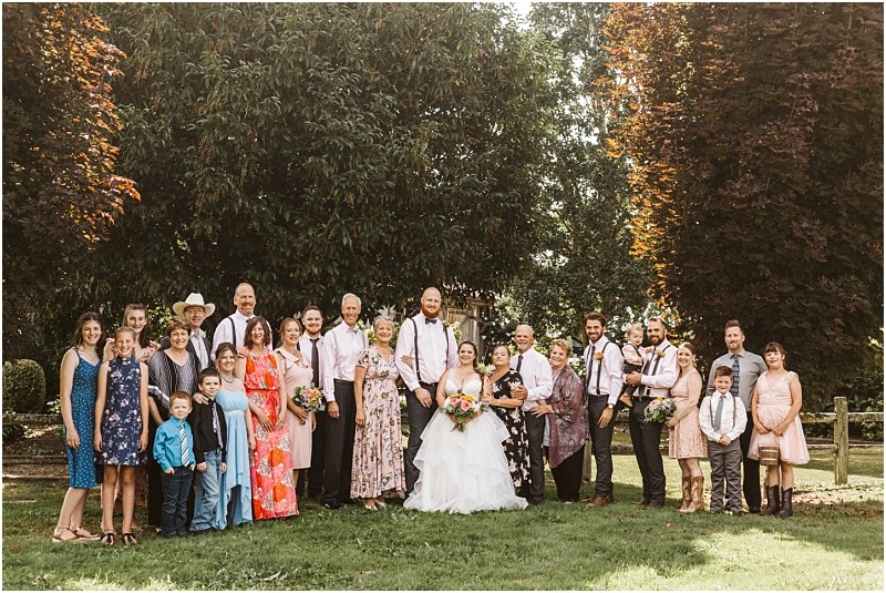 Premier Snohomish & Seattle Wedding Photographer | GSquared Weddings Photography A large group poses outdoors for a Snohomish wedding photo. The bride and groom stand at the center, surrounded by family and friends in semi-formal attire, framed by trees and sunlight. A perfect Seattle engagement celebration. 47.9129° N, 122.0982° W | Serving Snohomish, Seattle , Orlando & Beyond