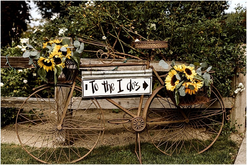 Premier Snohomish & Seattle Wedding Photographer | GSquared Weddings Photography A decorative vintage bicycle with baskets of sunflowers and greenery leans against a wooden fence, perfect for a Snohomish or Seattle wedding. A white sign on the bike reads "To the I dos" with an arrow pointing right. 47.9129° N, 122.0982° W | Serving Snohomish, Seattle , Orlando & Beyond