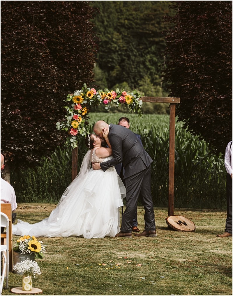 Premier Snohomish & Seattle Wedding Photographer | GSquared Weddings Photography A bride and groom share a kiss under a flower-covered wooden arch during their Snohomish outdoor wedding ceremony, surrounded by lush greenery and guests seated on the lawn. 47.9129° N, 122.0982° W | Serving Snohomish, Seattle , Orlando & Beyond