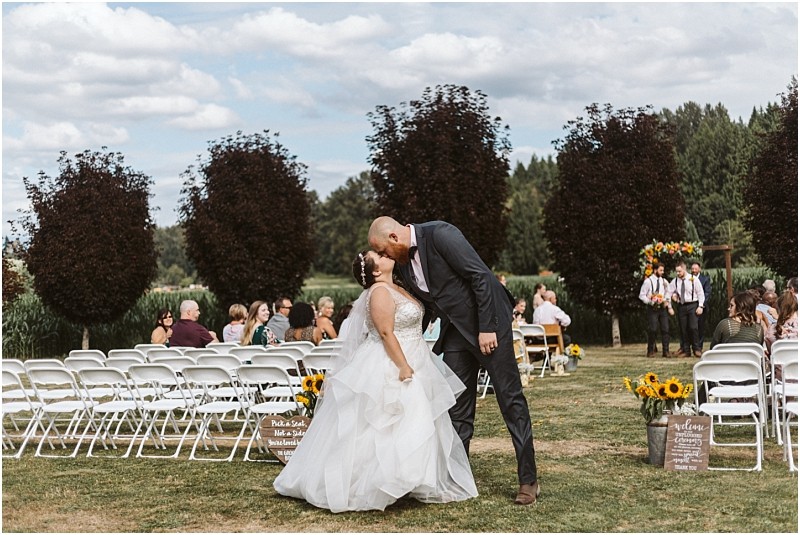 Premier Snohomish & Seattle Wedding Photographer | GSquared Weddings Photography A bride and groom share a kiss outdoors during their Snohomish wedding ceremony, surrounded by guests on white chairs and lush greenery, with sunflowers and trees in the background. 47.9129° N, 122.0982° W | Serving Snohomish, Seattle , Orlando & Beyond