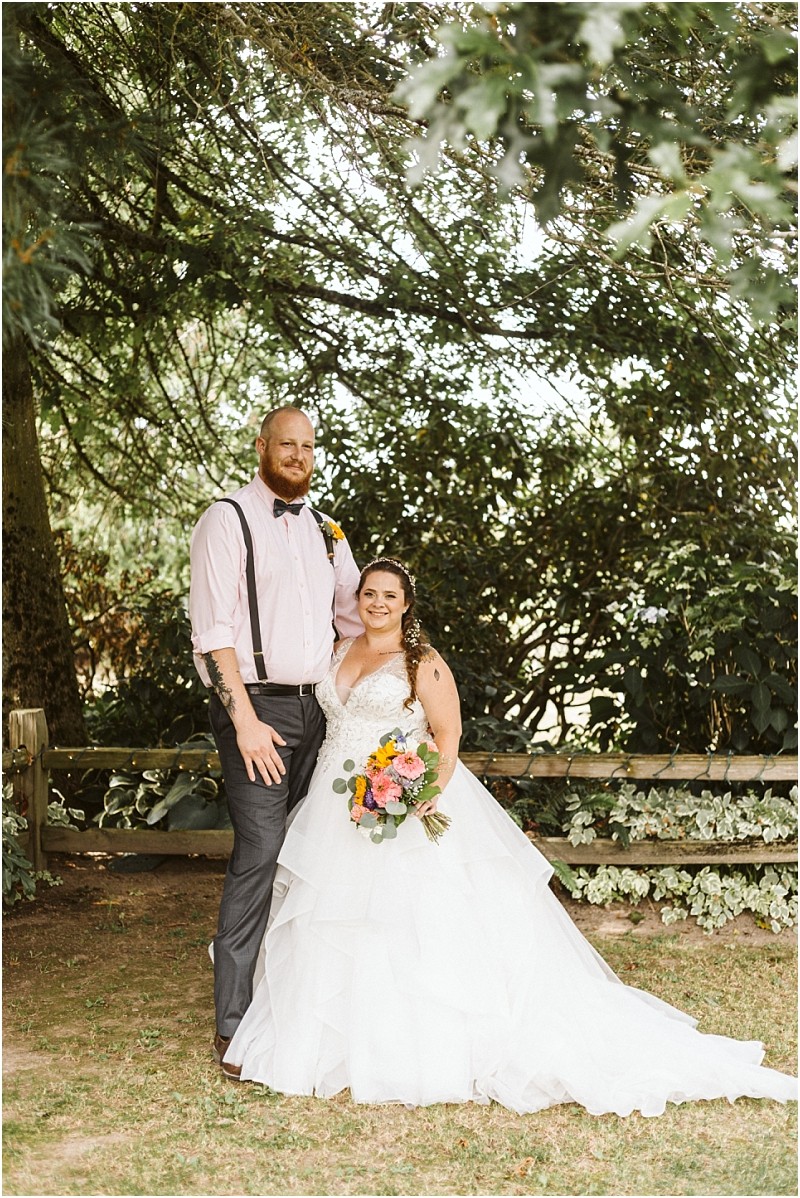 Premier Snohomish & Seattle Wedding Photographer | GSquared Weddings Photography A bride in a white gown holding a colorful bouquet stands beside a groom in dress pants, suspenders, and a light pink shirt. Smiling outdoors, they create unforgettable Snohomish Seattle wedding memories amid leafy trees and a wooden fence. 47.9129° N, 122.0982° W | Serving Snohomish, Seattle , Orlando & Beyond