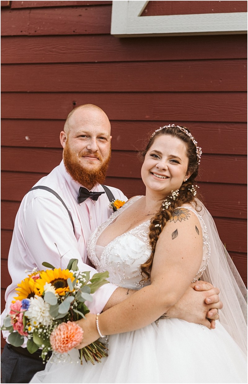 Premier Snohomish & Seattle Wedding Photographer | GSquared Weddings Photography A bride and groom stand smiling in front of a red wooden wall at their Snohomish wedding. The bride holds a colorful bouquet with sunflowers; the groom wears a light pink shirt, bow tie, and suspenders. 47.9129° N, 122.0982° W | Serving Snohomish, Seattle , Orlando & Beyond