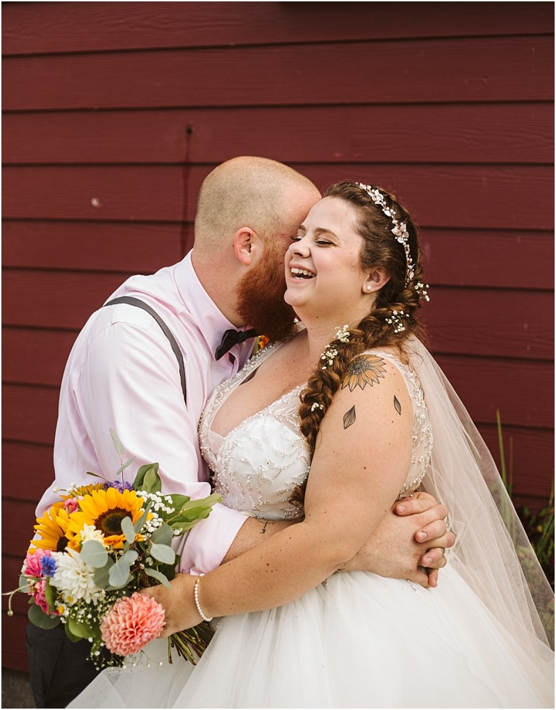 Premier Snohomish & Seattle Wedding Photographer | GSquared Weddings Photography A bride and groom embrace joyfully in front of a red wooden wall at their Snohomish Seattle wedding. The groom kisses the bride’s head as she smiles brightly, holding a colorful bouquet, both dressed in elegant wedding attire. 47.9129° N, 122.0982° W | Serving Snohomish, Seattle , Orlando & Beyond