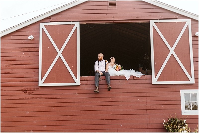 Premier Snohomish & Seattle Wedding Photographer | GSquared Weddings Photography A bride and groom sit in the open loft of a red barn with white trim, capturing a relaxed and happy moment. Perfect for a Snohomish Seattle wedding or engagement, the groom wears suspenders while the bride holds a colorful bouquet. 47.9129° N, 122.0982° W | Serving Snohomish, Seattle , Orlando & Beyond