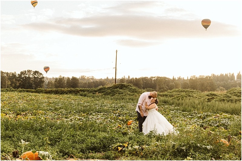 Premier Snohomish & Seattle Wedding Photographer | GSquared Weddings Photography A couple in wedding attire embraces and kisses in a lush green field with pumpkins, under a bright sky with hot air balloons floating in the distance—capturing the magic of a Snohomish Seattle wedding engagement. 47.9129° N, 122.0982° W | Serving Snohomish, Seattle , Orlando & Beyond