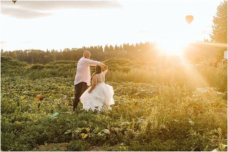 Premier Snohomish & Seattle Wedding Photographer | GSquared Weddings Photography A couple dances in a lush green field at sunset during their Snohomish Seattle wedding engagement, with the sun shining brightly behind them and a hot air balloon floating in the sky. The woman wears a white dress and the man wears a light shirt and dark pants. 47.9129° N, 122.0982° W | Serving Snohomish, Seattle , Orlando & Beyond