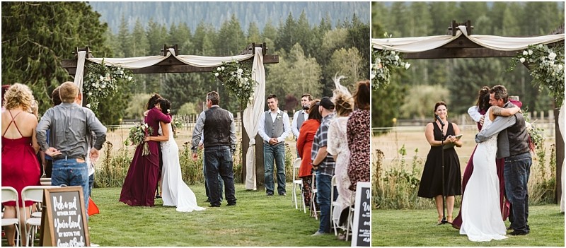 Premier Snohomish & Seattle Wedding Photographer | GSquared Weddings Photography Outdoor Snohomish wedding ceremony with a bride hugging a man under a flower-adorned arch. Guests watch from seated rows, some standing. Mountains and trees frame this beautiful Seattle engagement moment. 47.9129° N, 122.0982° W | Serving Snohomish, Seattle , Orlando & Beyond
