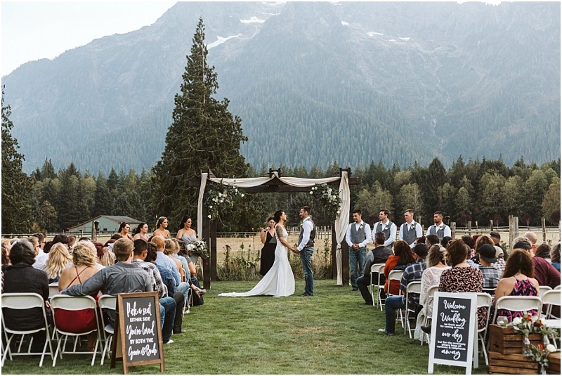 Premier Snohomish & Seattle Wedding Photographer | GSquared Weddings Photography A bride and groom stand under an arch during a romantic Snohomish outdoor wedding ceremony, surrounded by their wedding party and guests, with mountains and trees creating a stunning Seattle-area backdrop. 47.9129° N, 122.0982° W | Serving Snohomish, Seattle , Orlando & Beyond