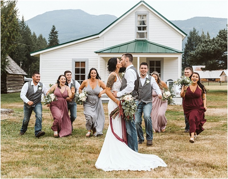 Premier Snohomish & Seattle Wedding Photographer | GSquared Weddings Photography A bride and groom kiss in front of a white house, surrounded by their joyful wedding party running toward them, all smiling and holding bouquets, with a stunning Snohomish mountain backdrop—a perfect Seattle wedding moment. 47.9129° N, 122.0982° W | Serving Snohomish, Seattle , Orlando & Beyond