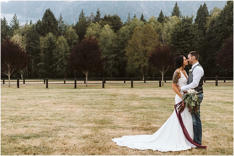 Premier Snohomish & Seattle Wedding Photographer | GSquared Weddings Photography A bride and groom stand close together, kissing and holding a bouquet in a grassy Snohomish field with trees and mountains in the background. The bride wears a white gown with a long train, capturing the romance of their Seattle wedding engagement. 47.9129° N, 122.0982° W | Serving Snohomish, Seattle , Orlando & Beyond