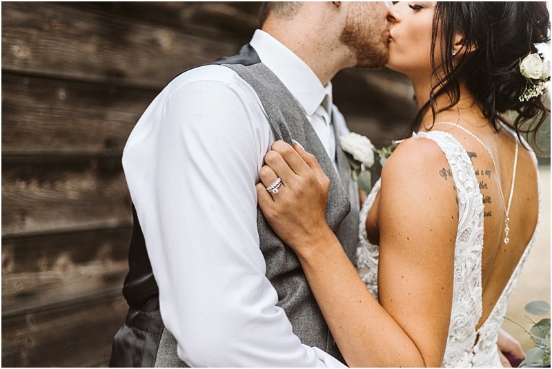 Premier Snohomish & Seattle Wedding Photographer | GSquared Weddings Photography A bride and groom share a kiss beside a wooden wall at their Snohomish wedding. The bride, in a white lace dress with her hair up and a flower in it, shows off her wedding ring while holding the groom’s shoulder. 47.9129° N, 122.0982° W | Serving Snohomish, Seattle , Orlando & Beyond
