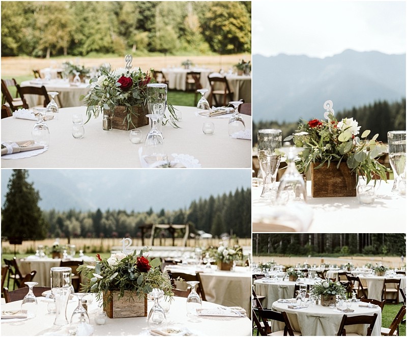 Premier Snohomish & Seattle Wedding Photographer | GSquared Weddings Photography Tables set outdoors with cream tablecloths, wooden chairs, and rustic centerpieces of greenery and red flowers in wooden boxes. Mountains, trees, and an open field create a scenic Snohomish wedding backdrop under a bright sky. 47.9129° N, 122.0982° W | Serving Snohomish, Seattle , Orlando & Beyond