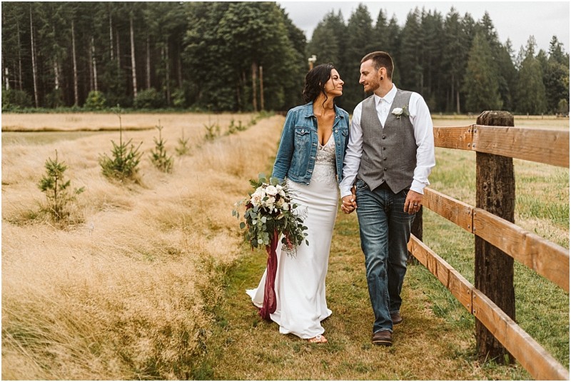 Premier Snohomish & Seattle Wedding Photographer | GSquared Weddings Photography A bride and groom walk hand in hand along a wooden fence in a grassy field at their Snohomish wedding. The bride wears a white dress and denim jacket, holding her bouquet, while the groom wears jeans, a vest, and white shirt. Trees frame the scene. 47.9129° N, 122.0982° W | Serving Snohomish, Seattle , Orlando & Beyond