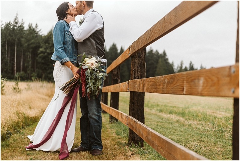Premier Snohomish & Seattle Wedding Photographer | GSquared Weddings Photography A bride in a white dress and denim jacket kisses her groom in a vest and jeans beside a wooden fence in a grassy field, holding a bouquet with burgundy ribbons. Forest trees hint at the rustic charm of a Snohomish Seattle wedding. 47.9129° N, 122.0982° W | Serving Snohomish, Seattle , Orlando & Beyond