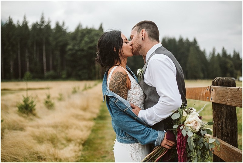 Premier Snohomish & Seattle Wedding Photographer | GSquared Weddings Photography A bride in a white dress and denim jacket kisses her groom in a vest and tie beside a wooden fence, holding a bouquet, during their Snohomish wedding, with a grassy field and trees in the background. 47.9129° N, 122.0982° W | Serving Snohomish, Seattle , Orlando & Beyond