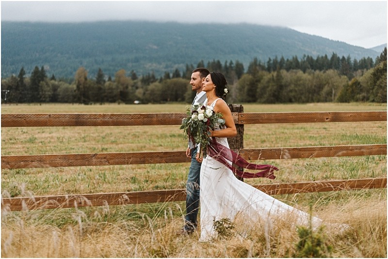 Premier Snohomish & Seattle Wedding Photographer | GSquared Weddings Photography A bride and groom walk side by side along a wooden fence in a grassy field near Snohomish, with mountains and trees in the background. The bride holds a bouquet and wears a flowing white dress; the groom is in jeans and a vest. 47.9129° N, 122.0982° W | Serving Snohomish, Seattle , Orlando & Beyond