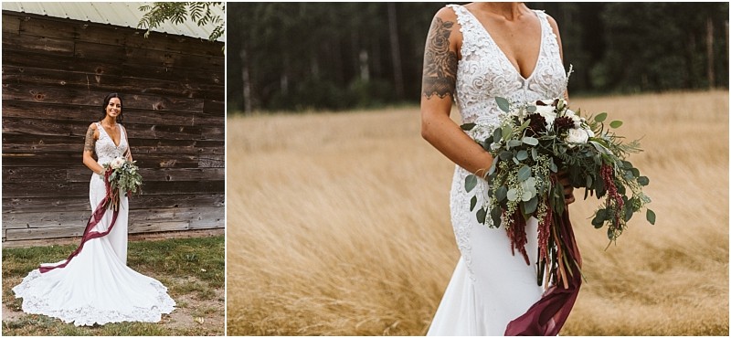 Premier Snohomish & Seattle Wedding Photographer | GSquared Weddings Photography A bride in a fitted white lace dress with a long train stands outdoors at a rustic Snohomish venue, holding a large bouquet of greenery and white flowers with a burgundy ribbon. She smiles beside a wooden wall, her arm tattoos visible—perfect for any Seattle wedding inspiration. 47.9129° N, 122.0982° W | Serving Snohomish, Seattle , Orlando & Beyond