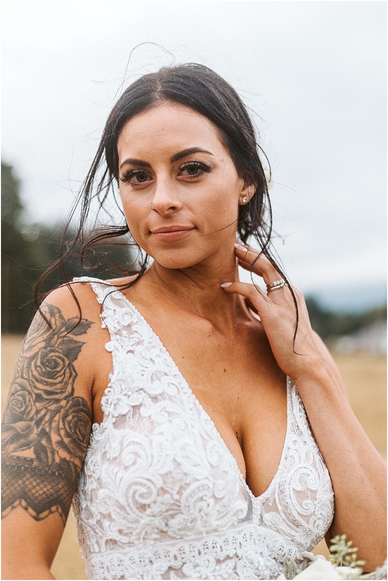 Premier Snohomish & Seattle Wedding Photographer | GSquared Weddings Photography A woman with long dark hair in a white lace dress stands outdoors near Snohomish, touching her neck. She has a floral tattoo on her upper arm and wears a ring, with a calm expression and a blurred natural background. 47.9129° N, 122.0982° W | Serving Snohomish, Seattle , Orlando & Beyond