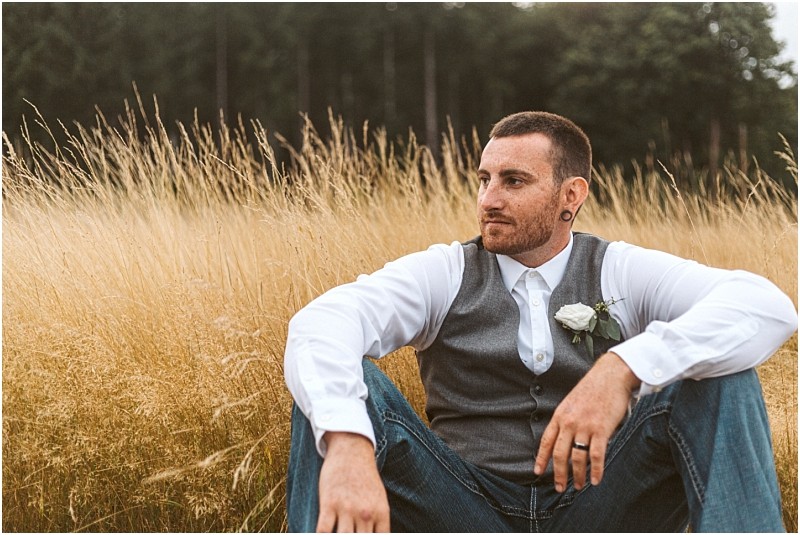 Premier Snohomish & Seattle Wedding Photographer | GSquared Weddings Photography A man with short hair and a beard sits in tall, golden grass at a Snohomish Seattle wedding engagement. He wears a white shirt, gray vest, jeans, and boutonniere, looking thoughtfully into the distance with trees in the background. 47.9129° N, 122.0982° W | Serving Snohomish, Seattle , Orlando & Beyond