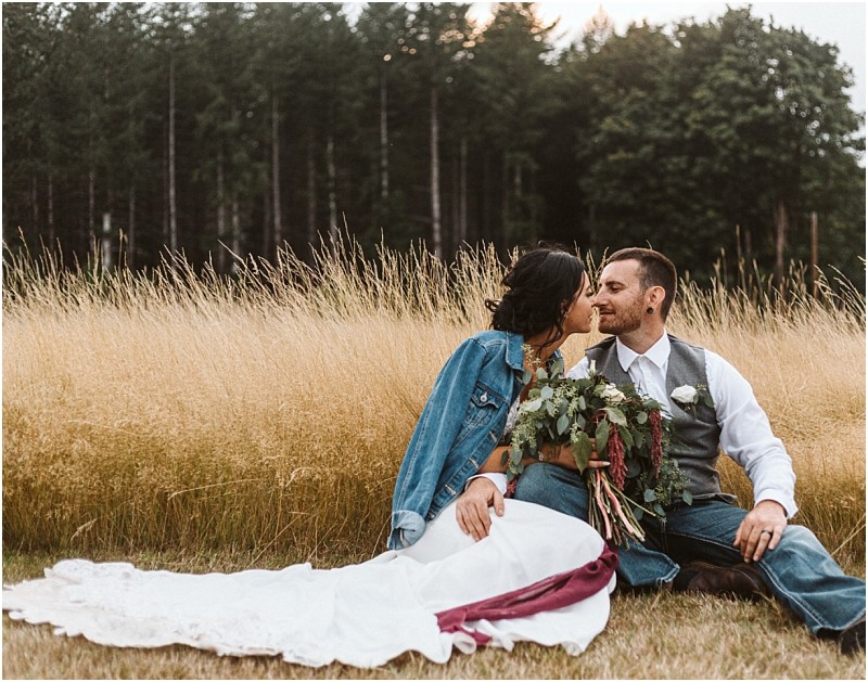 Premier Snohomish & Seattle Wedding Photographer | GSquared Weddings Photography A bride in a white dress and denim jacket sits in tall grass, holding a bouquet, and kisses her groom in a vest and jeans during their Snohomish wedding. Trees frame the scene under a cloudy Seattle sky. 47.9129° N, 122.0982° W | Serving Snohomish, Seattle , Orlando & Beyond