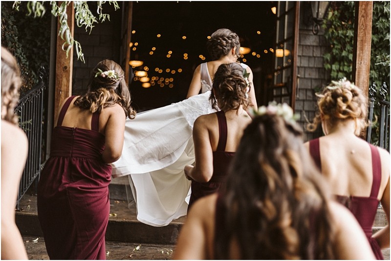 Premier Snohomish & Seattle Wedding Photographer | GSquared Weddings Photography Five women in burgundy dresses, likely bridesmaids, walk up steps into a Snohomish wedding venue. One woman lifts the train of a bride’s white dress as they enter, warmly lit by string lights inside. Perfect for a Seattle engagement celebration. 47.9129° N, 122.0982° W | Serving Snohomish, Seattle , Orlando & Beyond