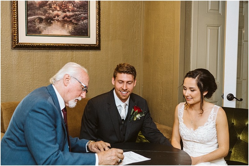 Premier Snohomish & Seattle Wedding Photographer | GSquared Weddings Photography A man in a blue suit signs a document at a table with a smiling bride in a white dress and groom in a dark suit, both seated and holding hands, during their Snohomish Seattle wedding. A framed painting hangs on the wall behind them. 47.9129° N, 122.0982° W | Serving Snohomish, Seattle , Orlando & Beyond
