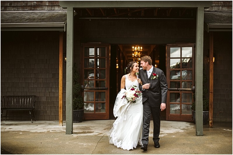 Premier Snohomish & Seattle Wedding Photographer | GSquared Weddings Photography A bride and groom, smiling and holding hands, walk out of a rustic building with wooden doors after their Snohomish wedding. The bride in a white gown holds a bouquet; the groom wears a dark suit and boutonniere. 47.9129° N, 122.0982° W | Serving Snohomish, Seattle , Orlando & Beyond