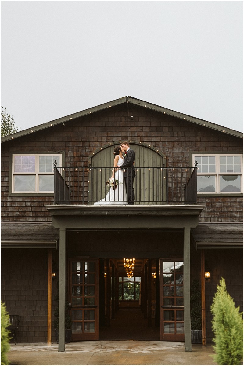 Premier Snohomish & Seattle Wedding Photographer | GSquared Weddings Photography A bride and groom stand close together on a balcony outside a rustic, dark-wood building in Snohomish, sharing a kiss in their wedding attire. String lights decorate the upper edge, capturing a romantic Seattle wedding moment. 47.9129° N, 122.0982° W | Serving Snohomish, Seattle , Orlando & Beyond