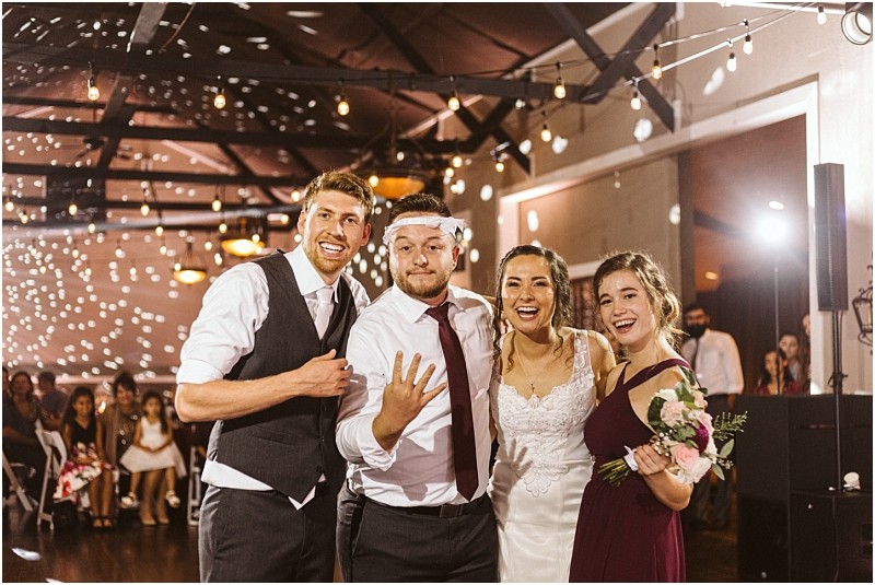 Premier Snohomish & Seattle Wedding Photographer | GSquared Weddings Photography Four people pose and smile at a Snohomish wedding reception: the groom with a garter on his head, the bride in white, a bridesmaid in burgundy holding flowers, and a groomsman. String lights and guests highlight this Seattle celebration. 47.9129° N, 122.0982° W | Serving Snohomish, Seattle , Orlando & Beyond