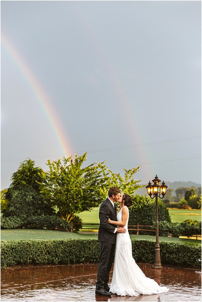 Premier Snohomish & Seattle Wedding Photographer | GSquared Weddings Photography A bride and groom embrace and kiss outdoors by a vintage lamp post, lush green trees, and a vivid double rainbow in the cloudy sky behind them—capturing a magical Snohomish Seattle wedding moment reflected on the wet ground. 47.9129° N, 122.0982° W | Serving Snohomish, Seattle , Orlando & Beyond