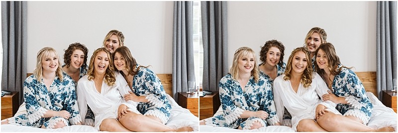 Premier Snohomish & Seattle Wedding Photographer | GSquared Weddings Photography Five women sit closely together on a bed, smiling and laughing in matching floral robes—except one in white—hinting at a Snohomish wedding bridal party getting ready. The bright background features curtains and a wooden headboard. 47.9129° N, 122.0982° W | Serving Snohomish, Seattle , Orlando & Beyond