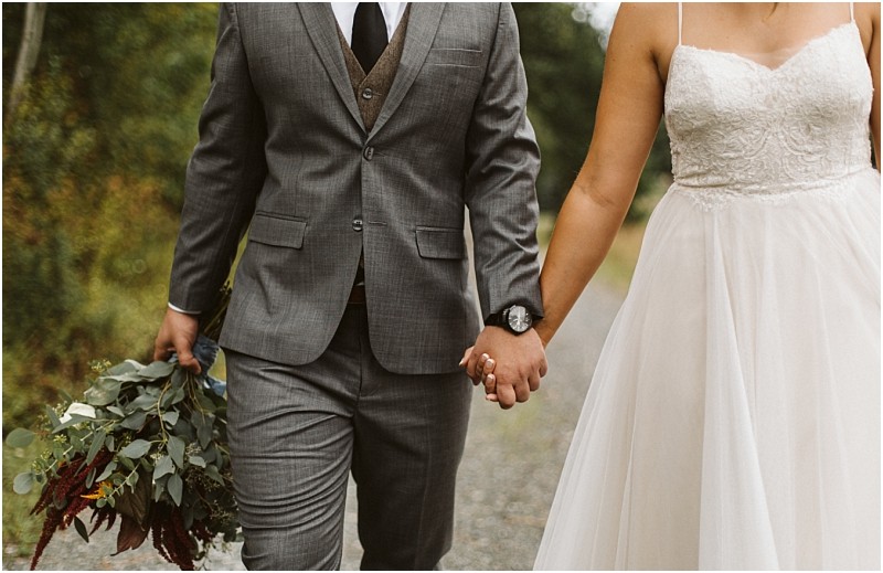 Premier Snohomish & Seattle Wedding Photographer | GSquared Weddings Photography A couple dressed in wedding attire holds hands while walking outdoors at a Snohomish Seattle wedding. The person on the left wears a gray suit and holds a bouquet; the person on the right wears a white lace wedding dress. 47.9129° N, 122.0982° W | Serving Snohomish, Seattle , Orlando & Beyond
