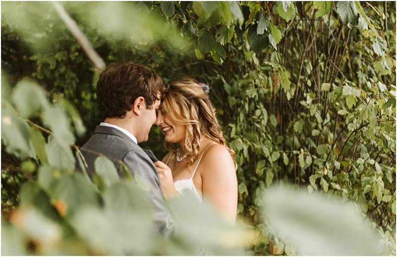 Premier Snohomish & Seattle Wedding Photographer | GSquared Weddings Photography A bride and groom stand close together, smiling and touching foreheads, partially hidden by green leafy branches in a beautiful Snohomish outdoor setting—capturing the romance of a Seattle wedding engagement. 47.9129° N, 122.0982° W | Serving Snohomish, Seattle , Orlando & Beyond