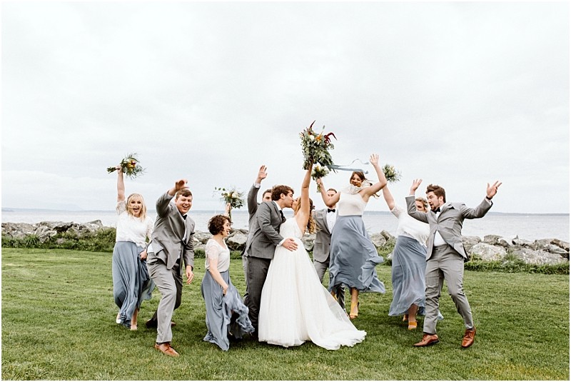 Premier Snohomish & Seattle Wedding Photographer | GSquared Weddings Photography A wedding party poses joyfully on a grassy lawn by the water at a stunning Snohomish Seattle wedding. The bride and groom stand center, surrounded by bridesmaids in blue dresses and groomsmen in gray suits, all celebrating with lifted bouquets. 47.9129° N, 122.0982° W | Serving Snohomish, Seattle , Orlando & Beyond