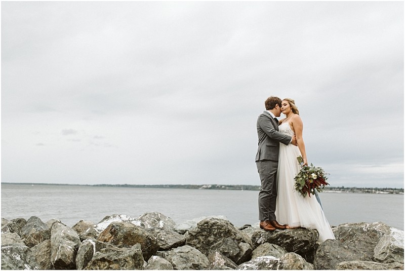 Premier Snohomish & Seattle Wedding Photographer | GSquared Weddings Photography A bride and groom stand on a rocky shore by the water near Seattle, embracing and kissing under a cloudy sky. The bride holds a bouquet and wears a white dress, while the groom, in a gray suit, shares this romantic Snohomish wedding moment. 47.9129° N, 122.0982° W | Serving Snohomish, Seattle , Orlando & Beyond