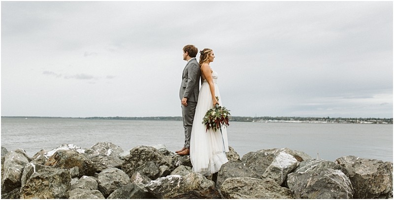 Premier Snohomish & Seattle Wedding Photographer | GSquared Weddings Photography A bride and groom stand back to back on large rocks by the water, facing opposite directions. The overcast sky and distant shoreline set a serene mood—perfect for a Snohomish Seattle wedding or engagement. The bride holds a bouquet of flowers. 47.9129° N, 122.0982° W | Serving Snohomish, Seattle , Orlando & Beyond