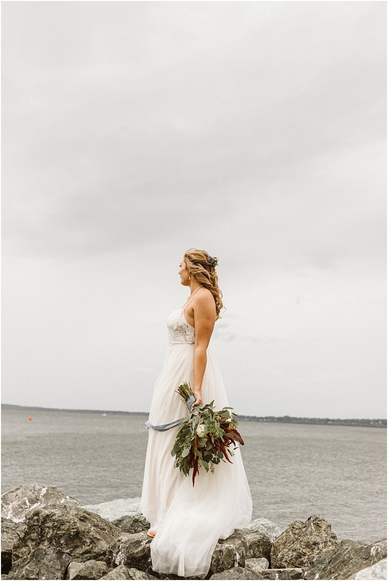 Premier Snohomish & Seattle Wedding Photographer | GSquared Weddings Photography A bride in a white gown stands on rocky ground by a body of water near Snohomish, holding a bouquet of greenery and flowers, gazing into the distance under a cloudy sky—capturing the magic of a Seattle wedding engagement. 47.9129° N, 122.0982° W | Serving Snohomish, Seattle , Orlando & Beyond
