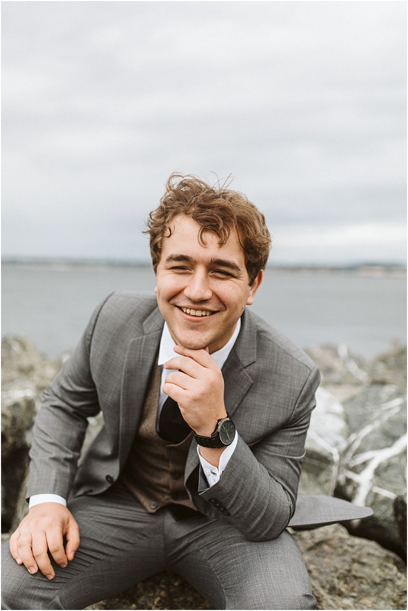 Premier Snohomish & Seattle Wedding Photographer | GSquared Weddings Photography A young man in a gray suit sits on rocks by the water, smiling warmly at the camera with his hand resting on his chin. The overcast sky and blurred shoreline create a serene backdrop for this Snohomish Seattle engagement moment. 47.9129° N, 122.0982° W | Serving Snohomish, Seattle , Orlando & Beyond